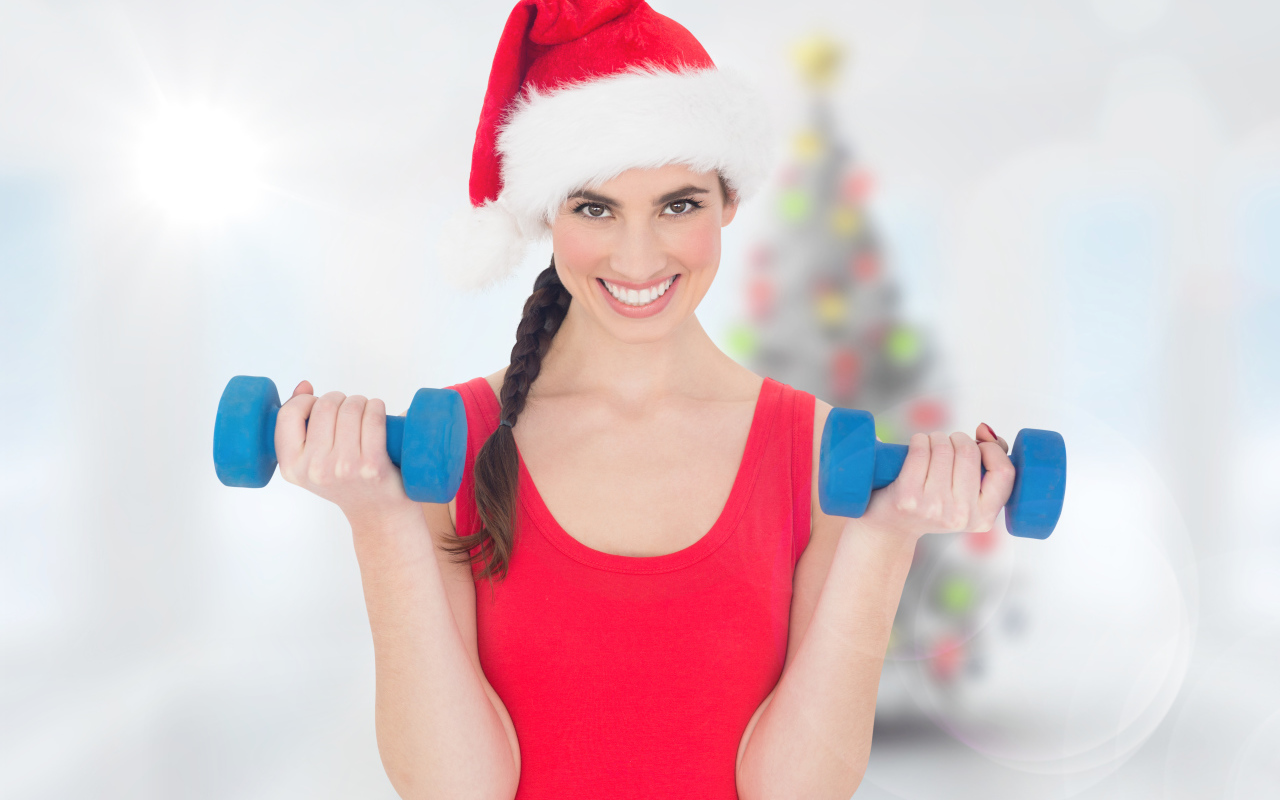 Красивая девушка в шапке Санта Клауса с гантелями в руках 