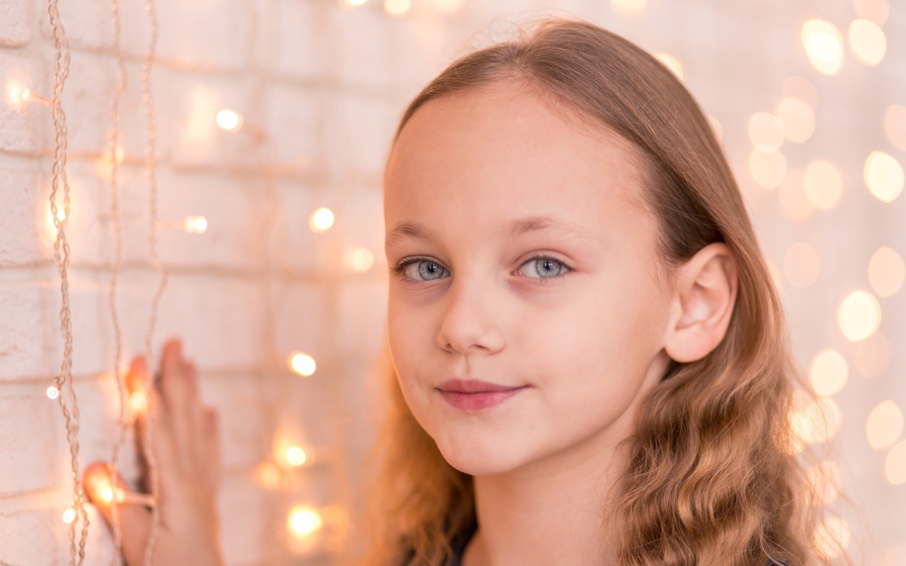 Красивая голубоглазая девочка стоит у стены с гирляндой