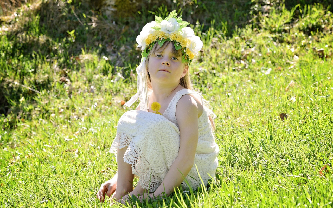 Маленькая девочка с венком на голове сидит на зеленой траве