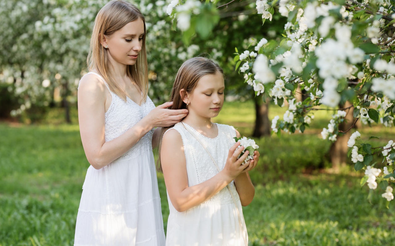 Мама заплетает дочке волосы у дерева