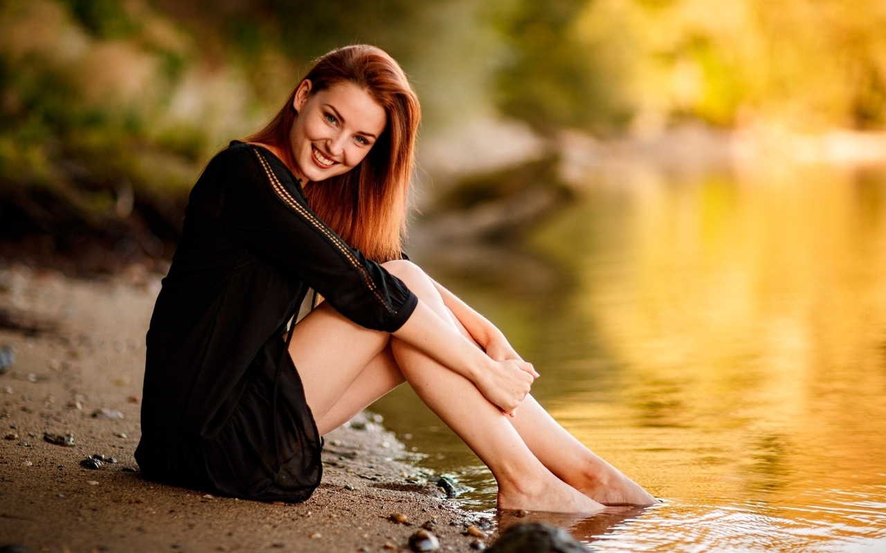 Улыбающаяся девушка в черном платье сидит у воды