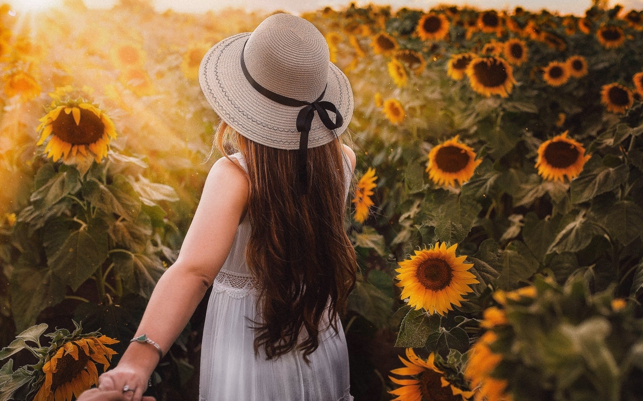 Девушка в белом платье и шляпе идет по полю с подсолнухами