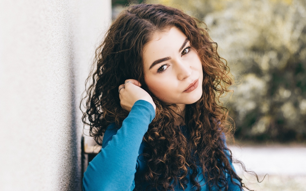 Кареглазая девушка с кудрявыми волосами стоит у стены