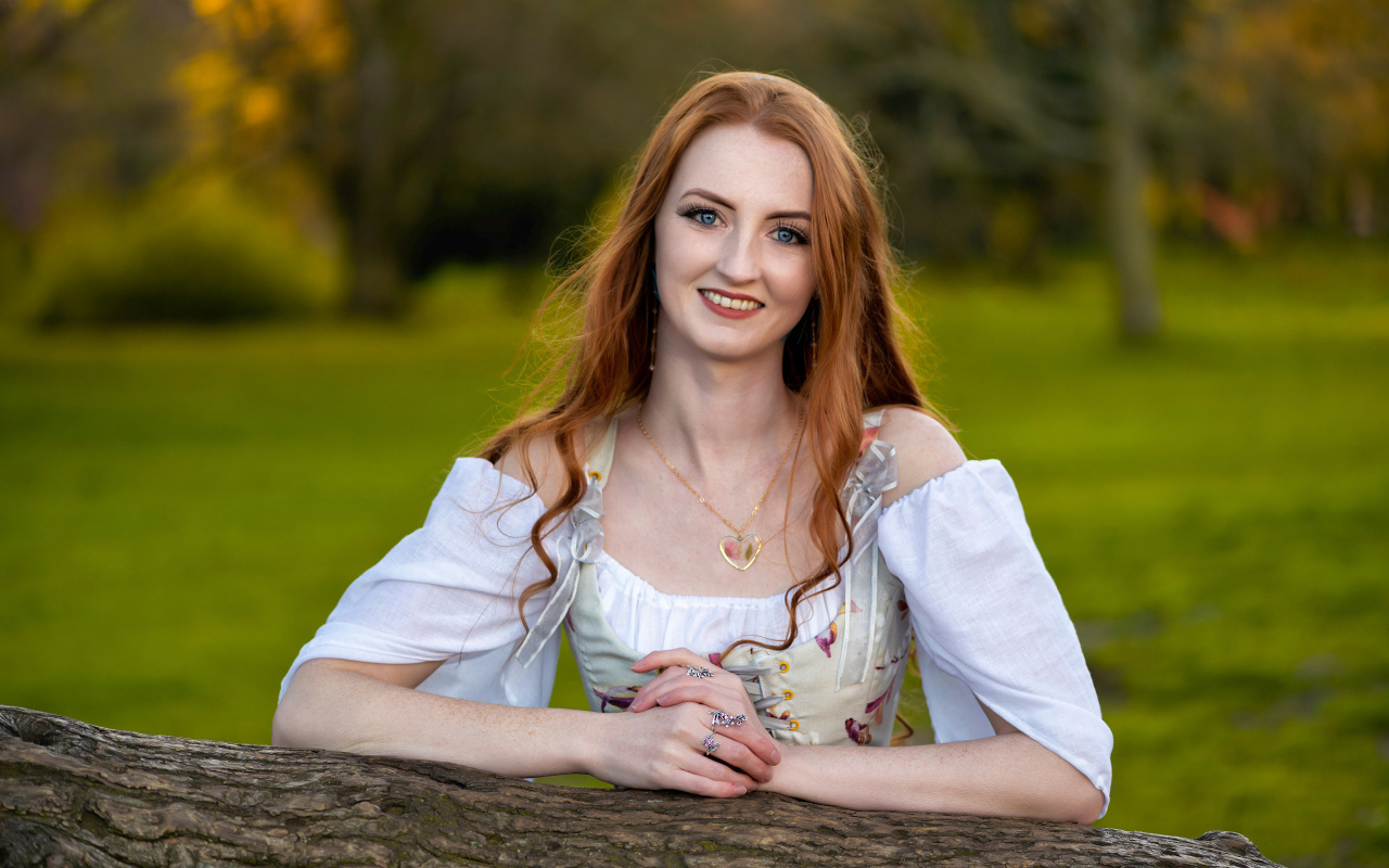 Улыбающаяся рыжеволосая девушка стоит у дерева