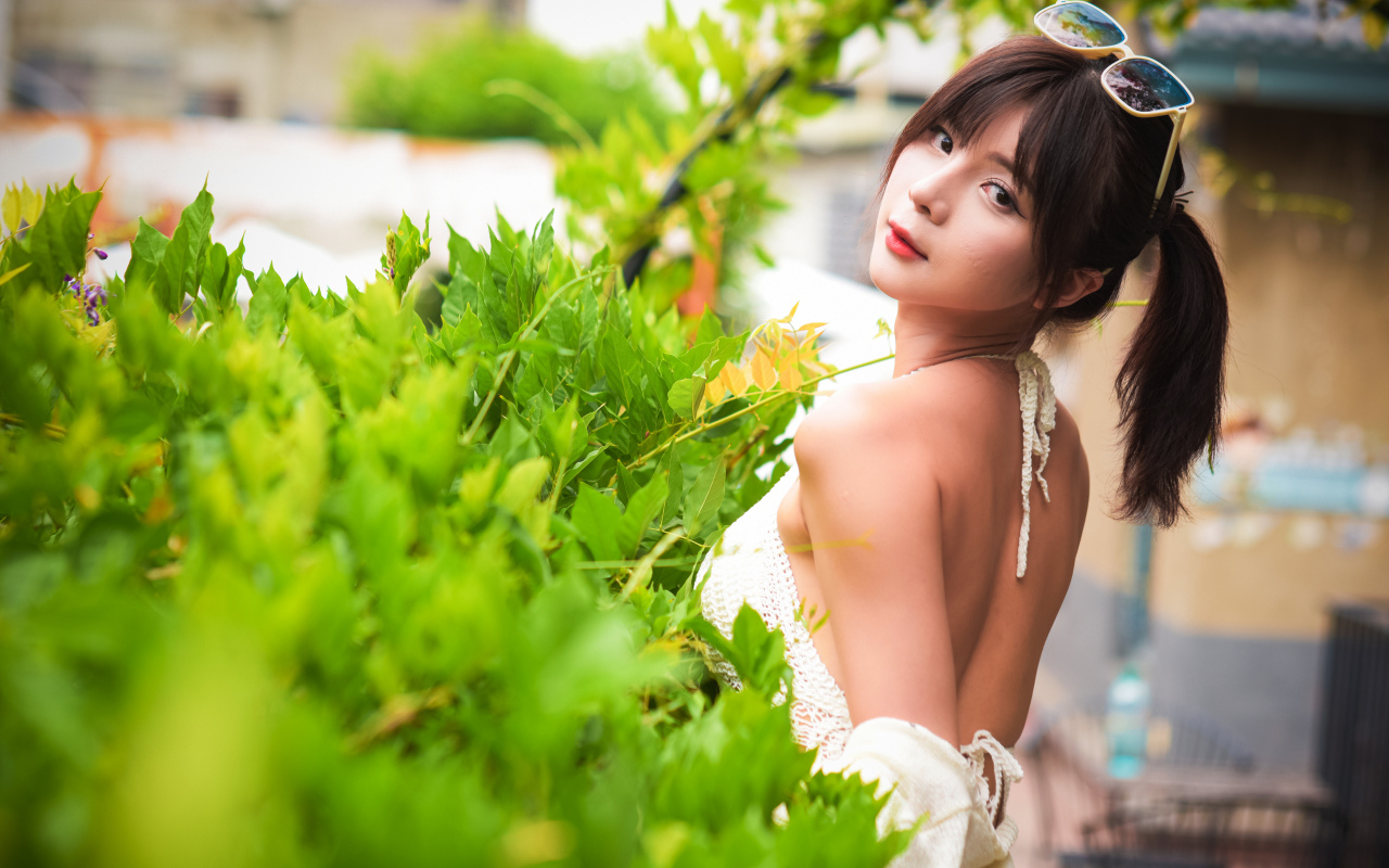 Девушка азиатка с очками на голове стоит у куста с зелеными листьями