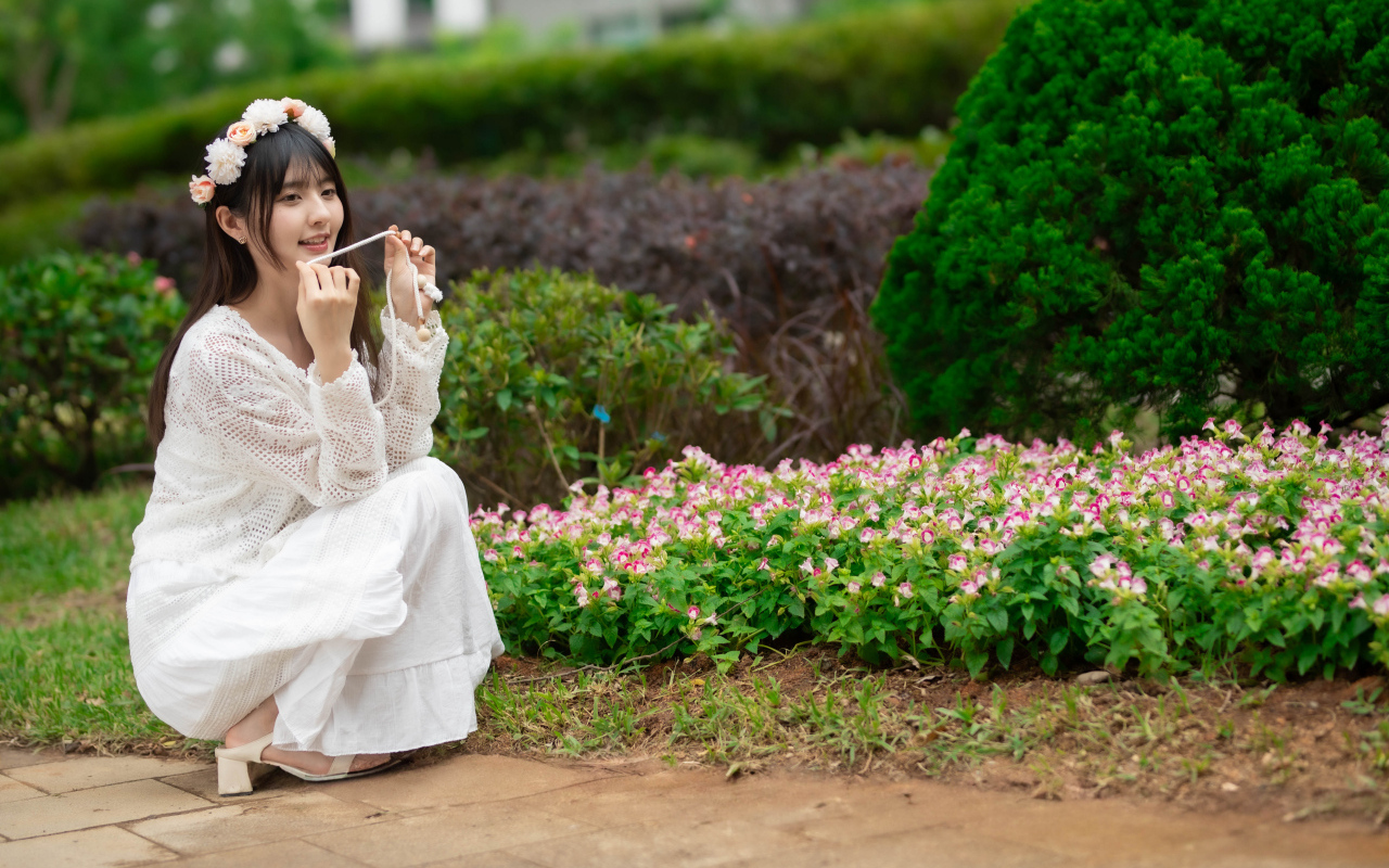 Милая азиатка в белом наряде сидит у клумбы с цветами