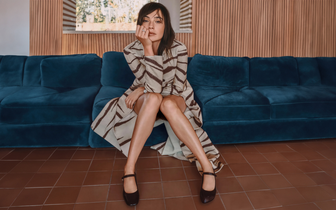 Актриса Галь Гадот в пальто сидит на диване