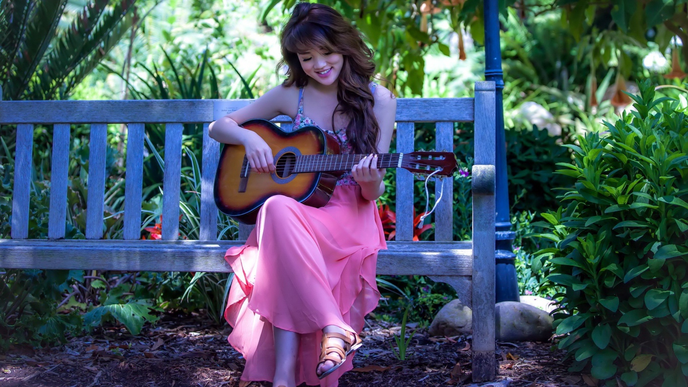 Девушка сидит на скамье в саду и играет на гитаре