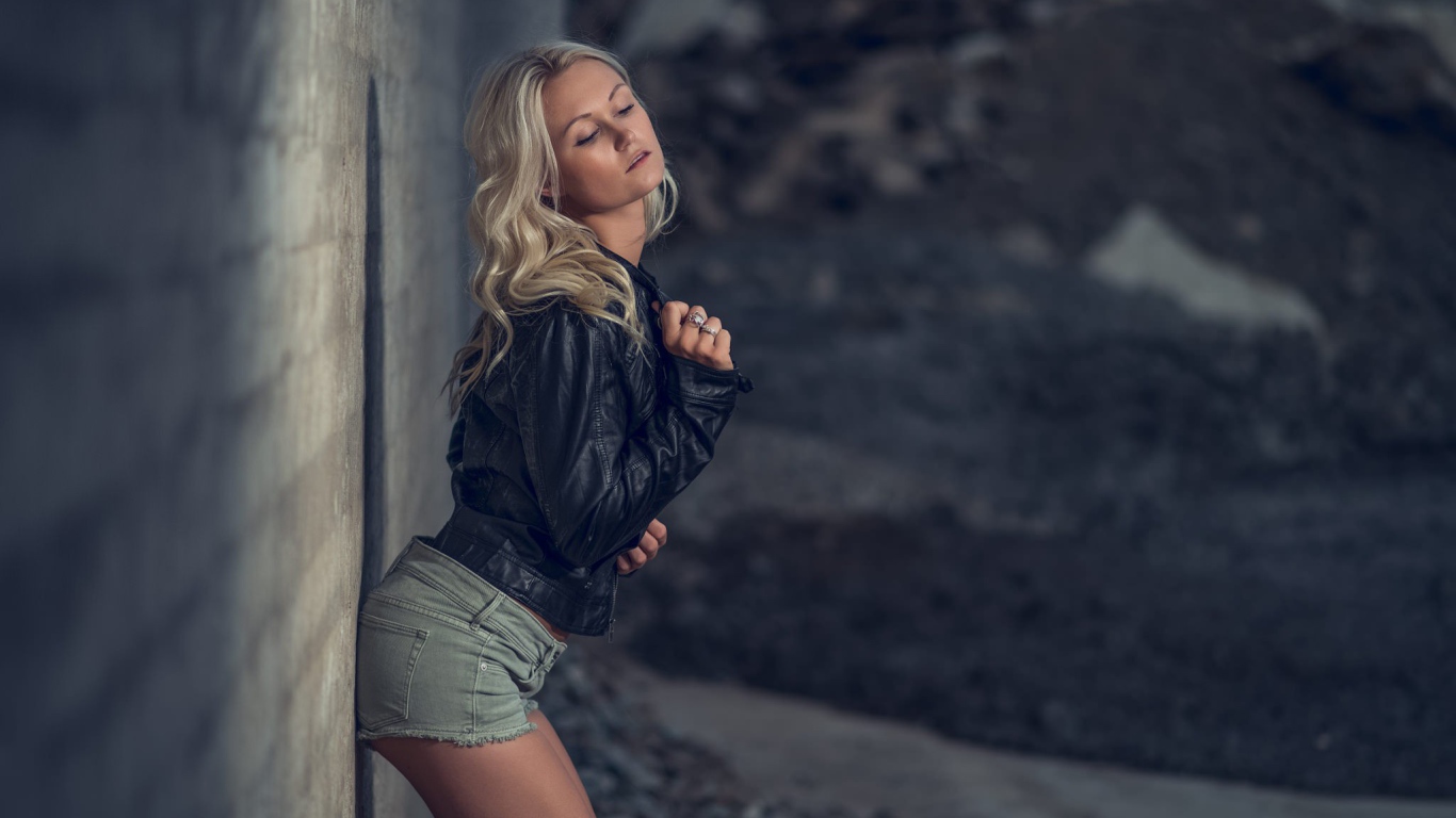 Блондинка в джинсовых шортах стоит у стены