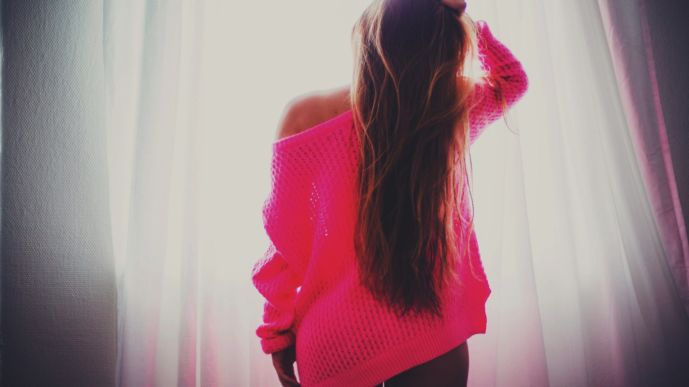 Брюнетка в розовой кофте стоит у окна