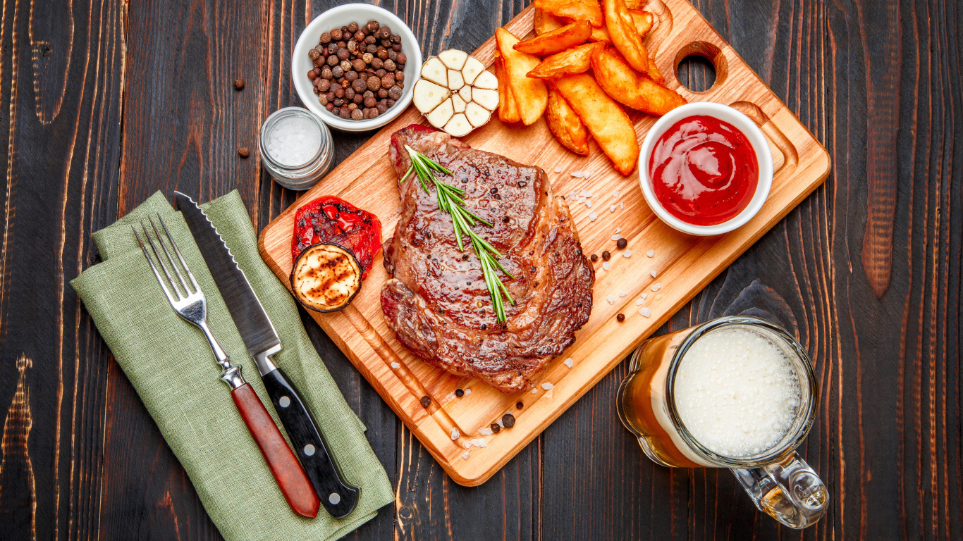 Сочный кусок мяса с картофелем фри, специями и бокалом пива на толе