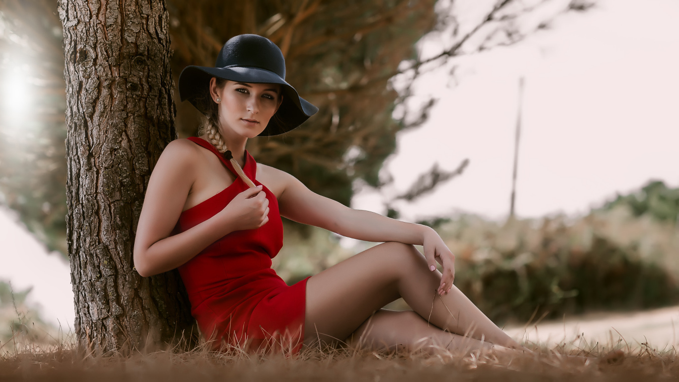 Красивая девушка в красном платье и черной шляпе сидит под деревом