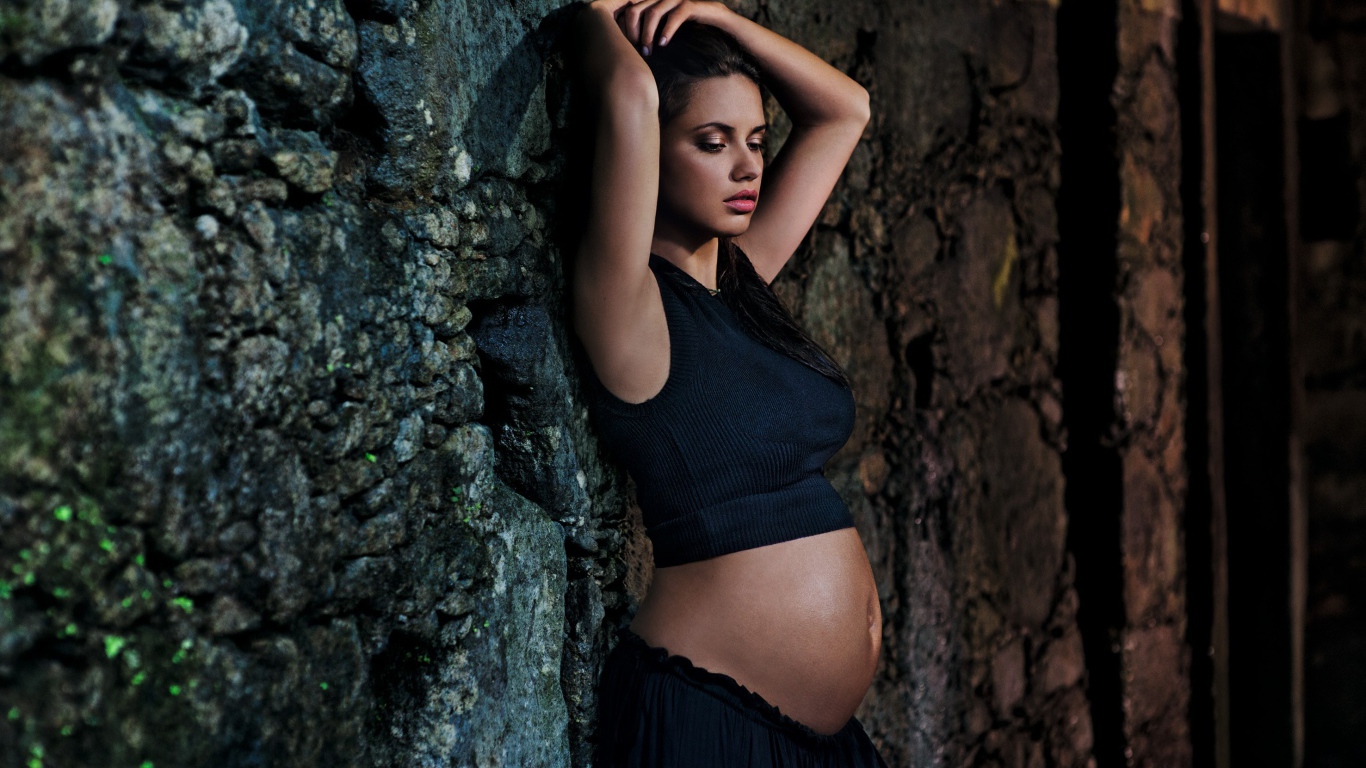 Беременная девушка в черном наряде у стены 