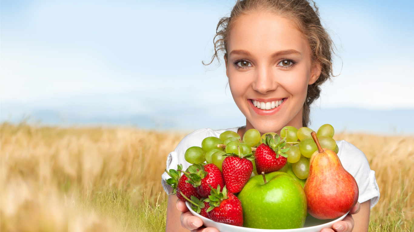 Улыбающаяся девушка шатенка с тарелкой с яблоками, виноградом, клубникой и грушей в руках