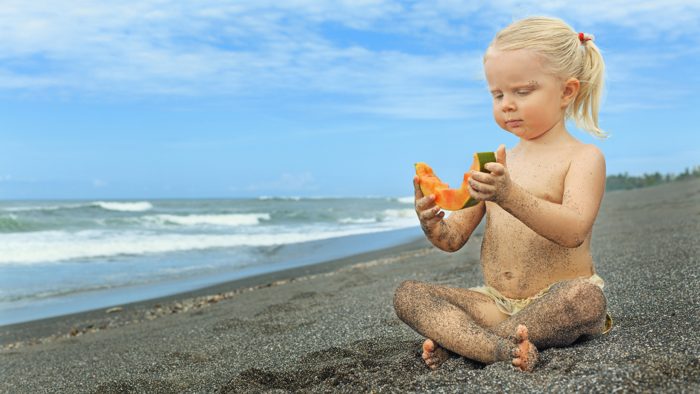 Маленькая девочка сидит на песке у моря с кусочком дыни