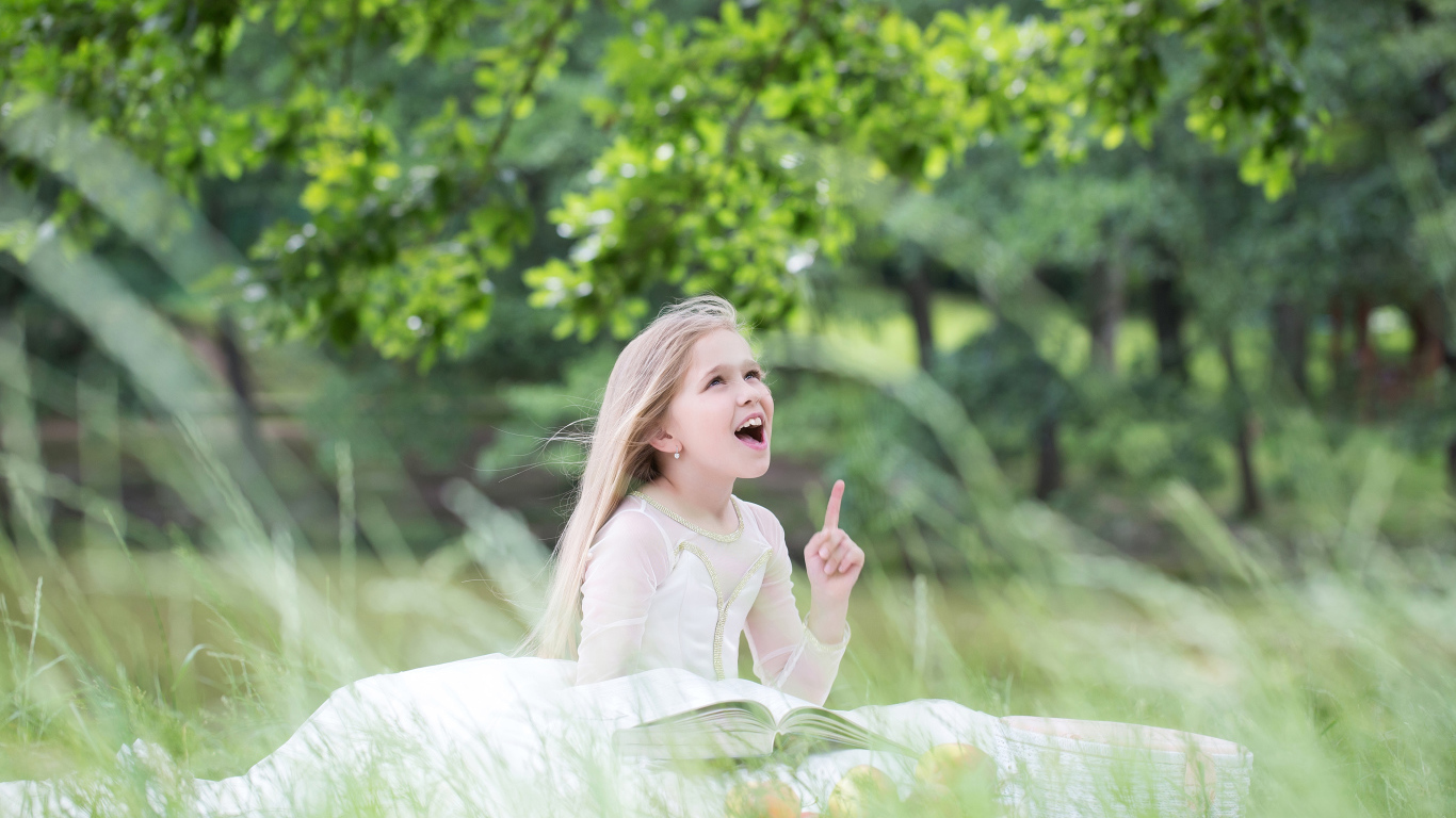 Красивая маленькая девочка в белом платье с книгой сидит на траве