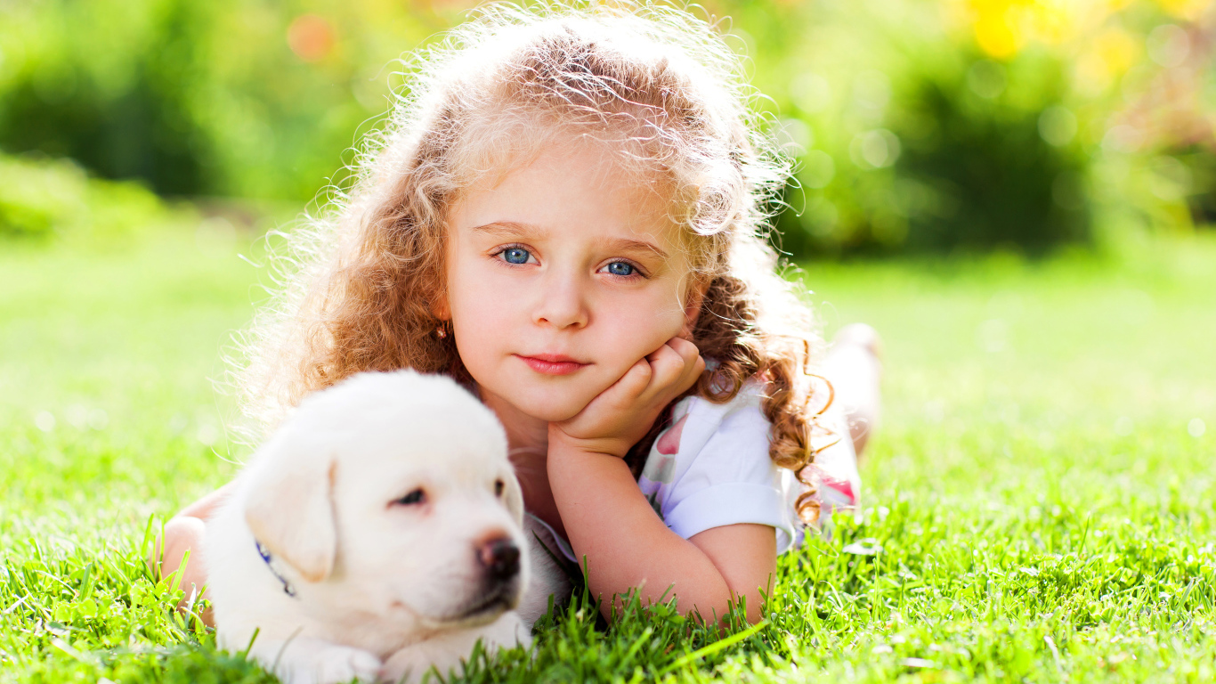 Маленькая голубоглазая девочка с щенком золотистого ретривера лежит на зеленой траве