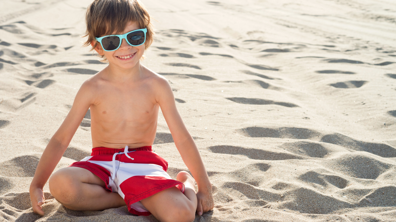 Маленький мальчик в очках сидит на песке на пляже