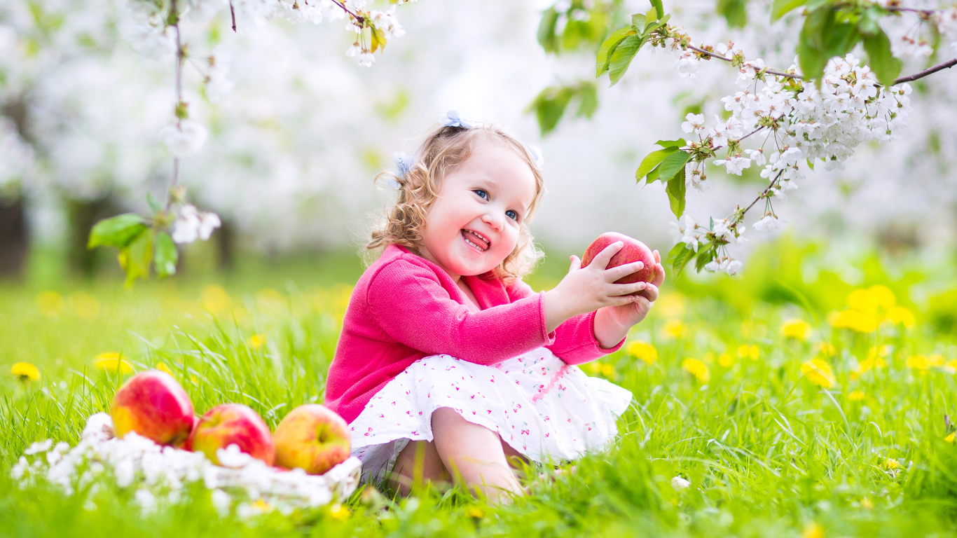 Маленькая улыбающаяся девочка сидит на зеленой траве с яблоками