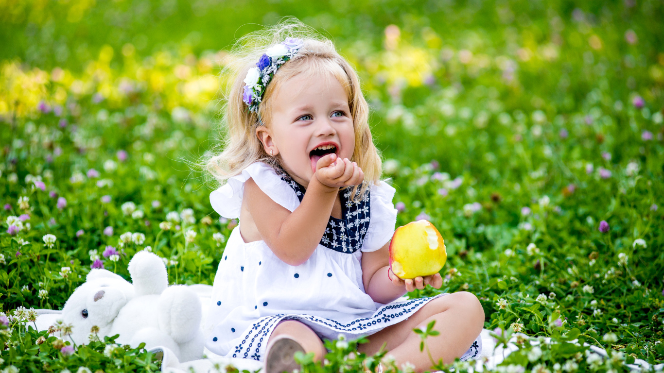 Маленькая улыбающаяся девочка с яблоком сидит на зеленой траве
