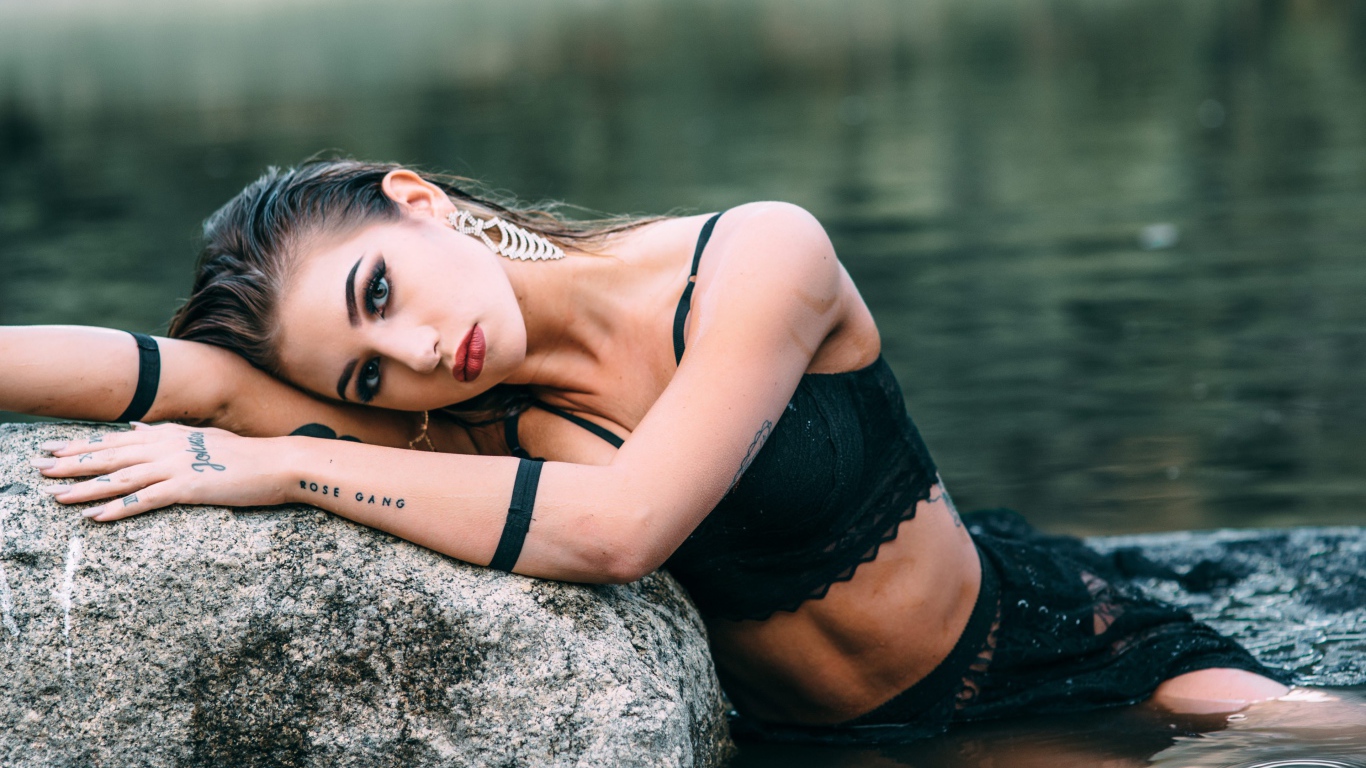 Молодая девушка с татуировками сидит у камня в воде
