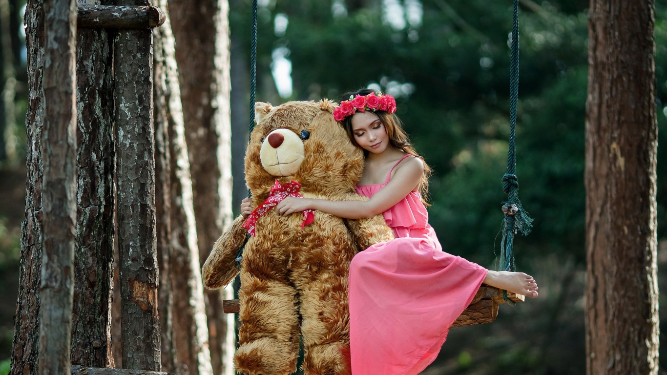 Красивая девушка азиатка в розовом платье сидит на качели с большим игрушечным медведем