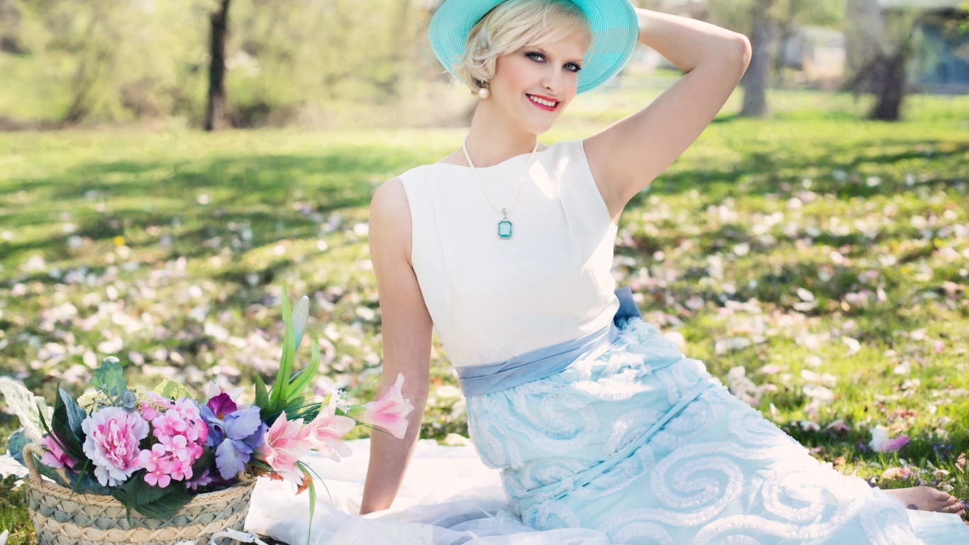 Красивая блондинка в голубой шляпке сидит в парке на траве