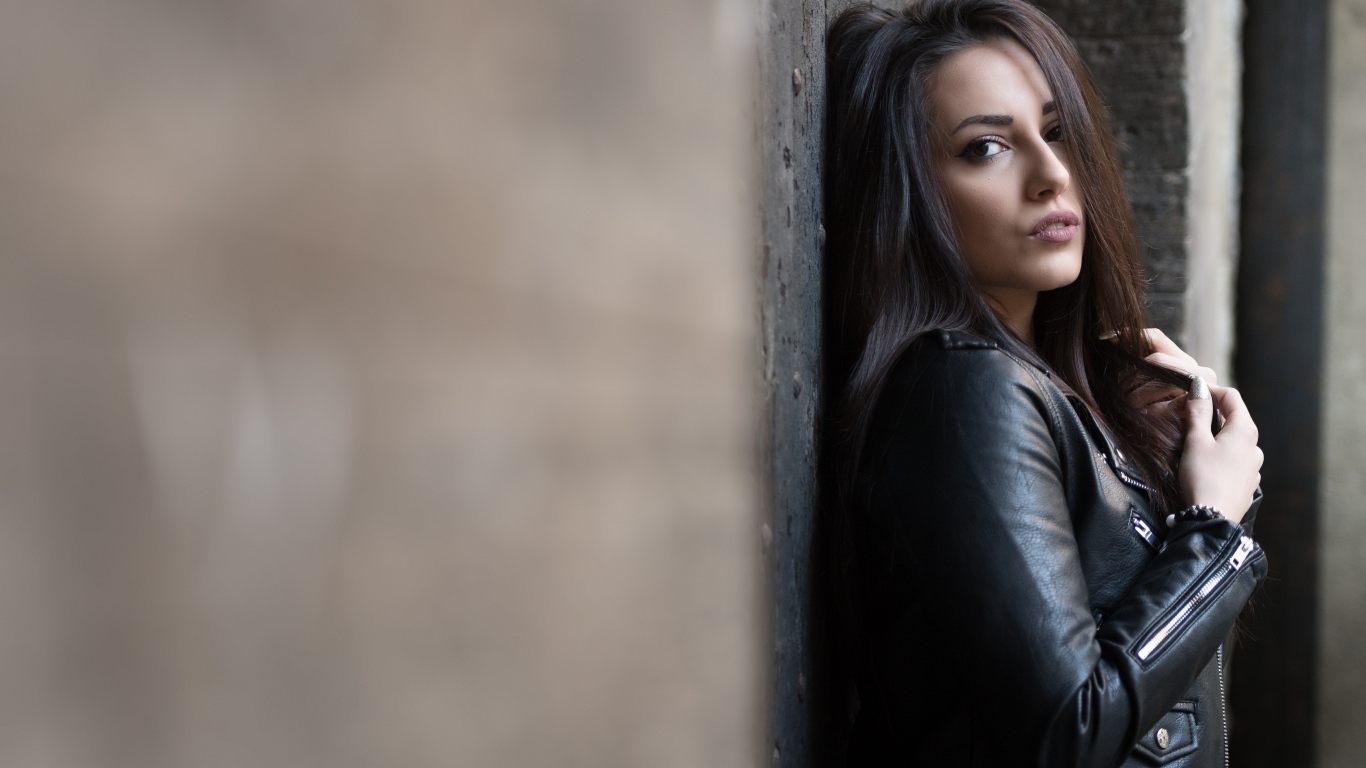 Красивая  брюнетка в кожаной куртке стоит у стены