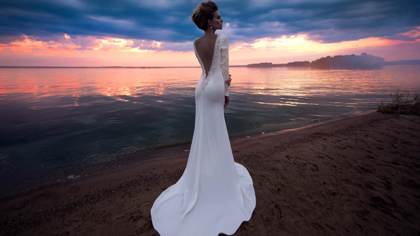 Красивая молодая девушка в свадебном платье у воды на закате 