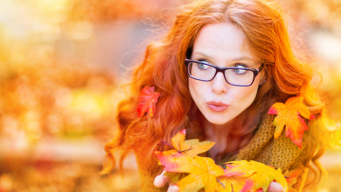Рыжеволосая девушка в очках держит в руках опавшие листья 