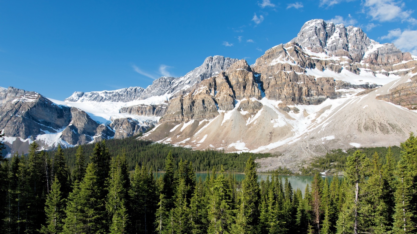 Заснеженные горы с озера у зеленого леса под голубым небом