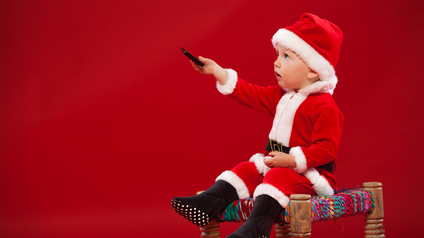 Маленький мальчик в костюме Санта Клауса сидит на стуле на красном фоне