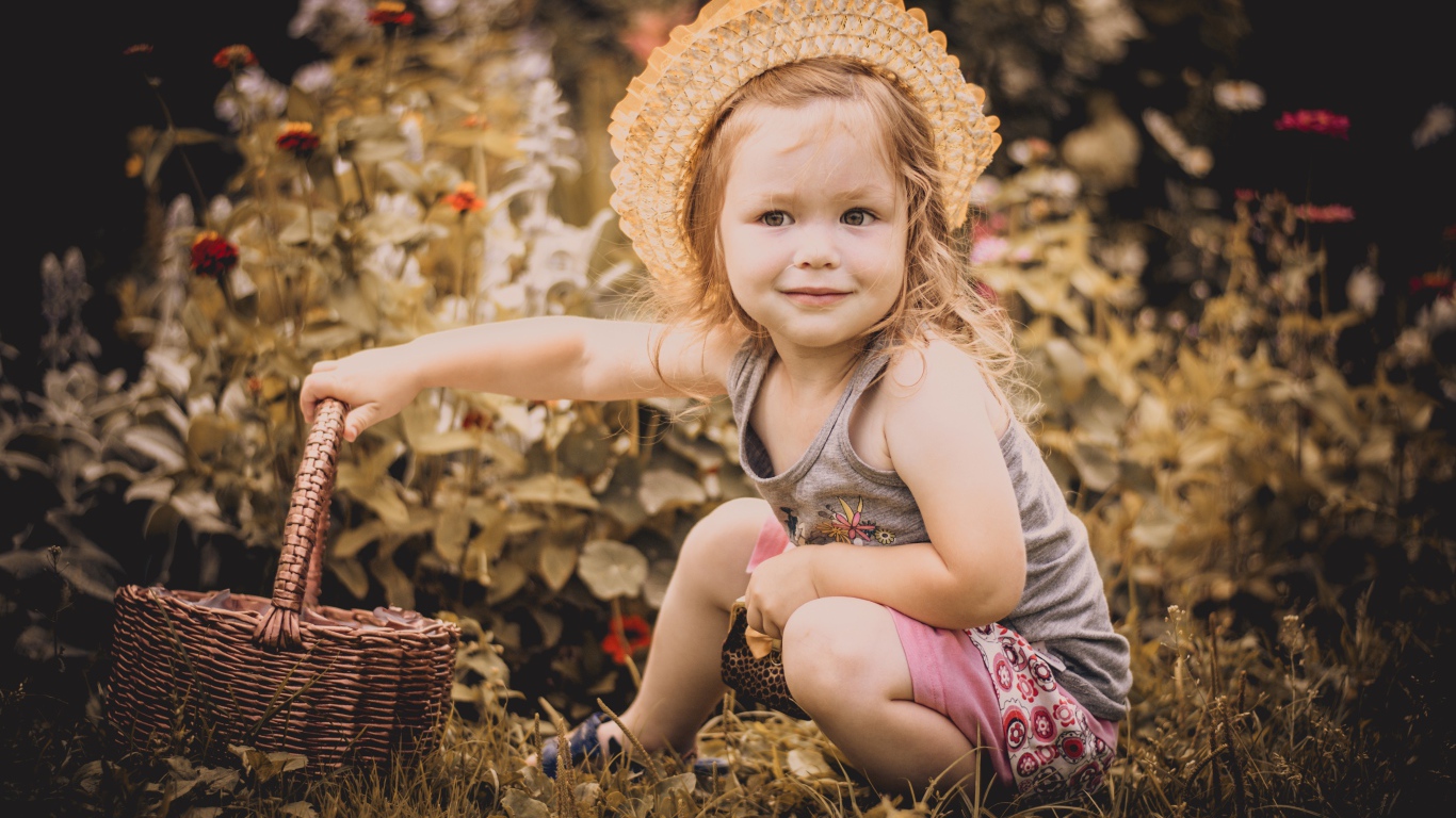 Маленькая девочка в шляпе сидит на траве с корзиной