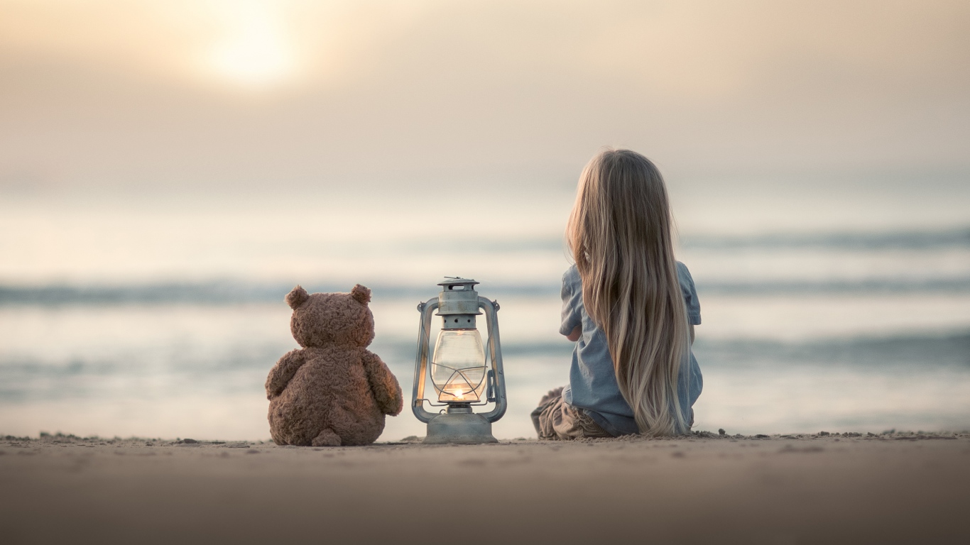 Маленькая девочка сидит на песке с плюшевым мишкой 