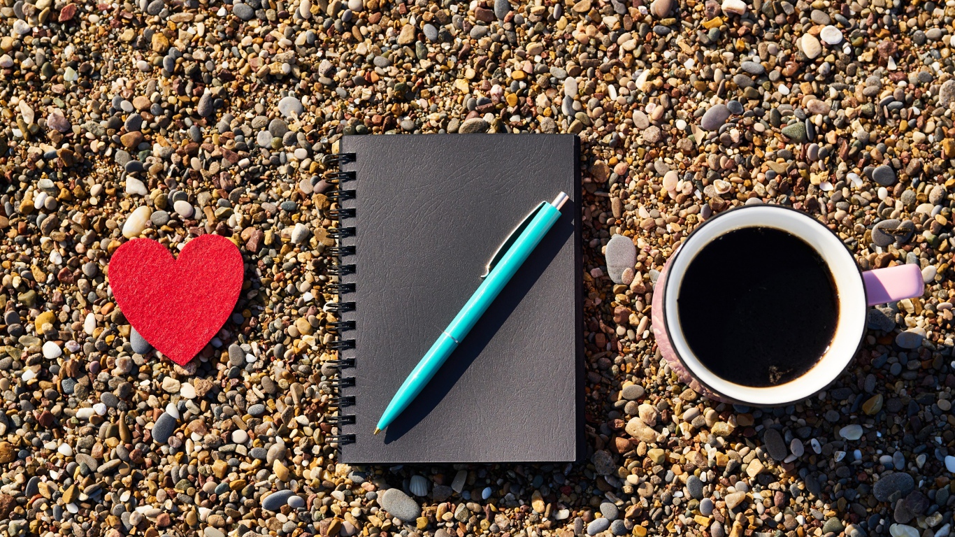 Блокнот с ручкой лет на мелкой гальке с чашкой кофе и красным сердцем 