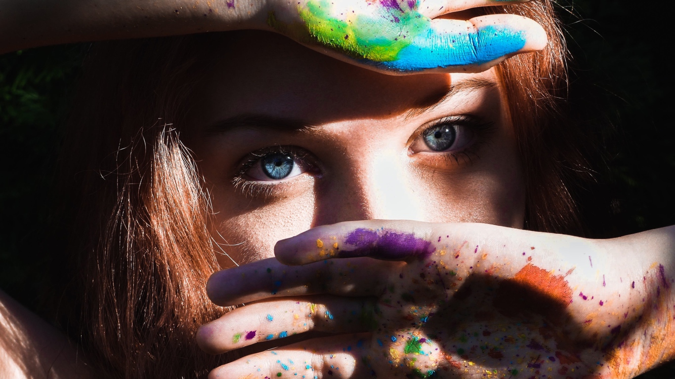 Красивая голубоглазая девушка с руками в разноцветной краске у лица