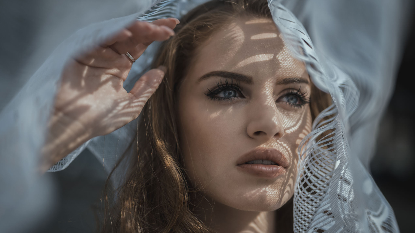 Голубоглазая девушка смотрит в окно с тюлью