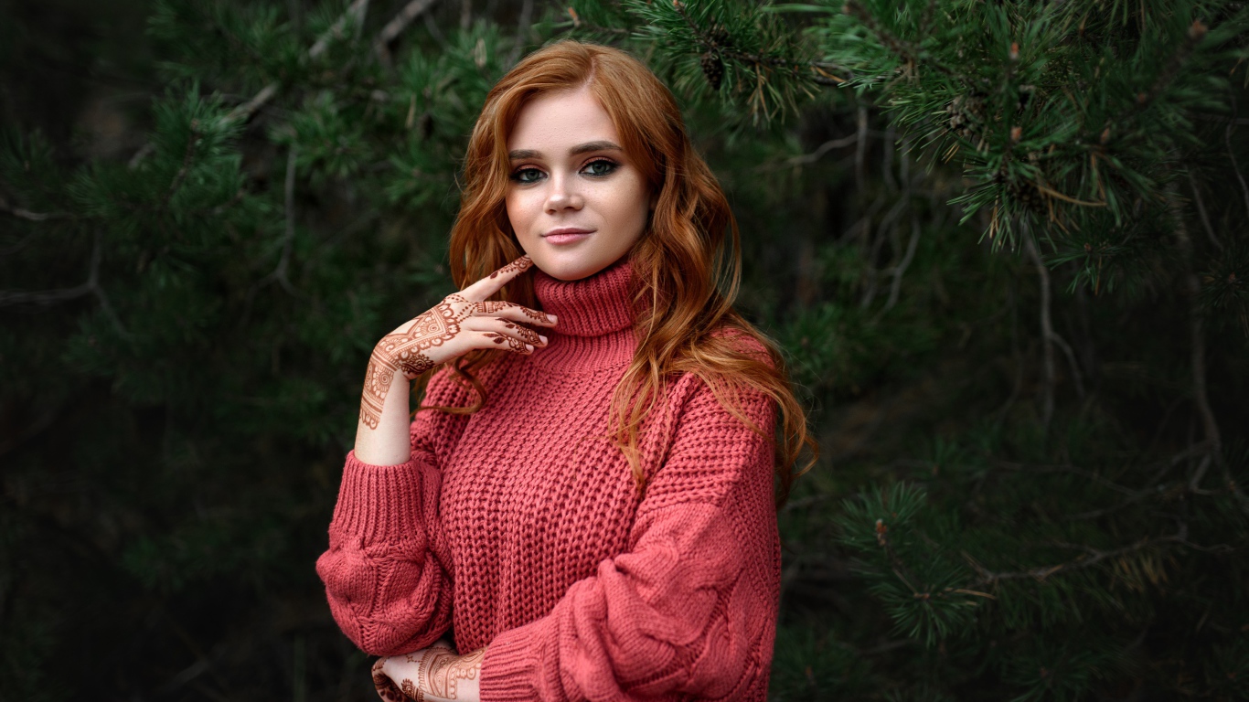 Рыжеволосая девушка в розовом свитере стоит у ели