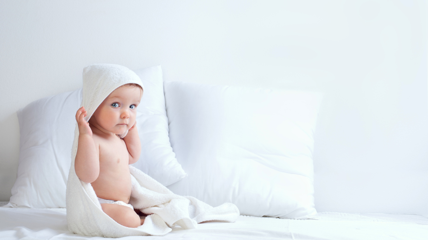 Маленький мальчик сидит в белом покрывале на кровати с подушками