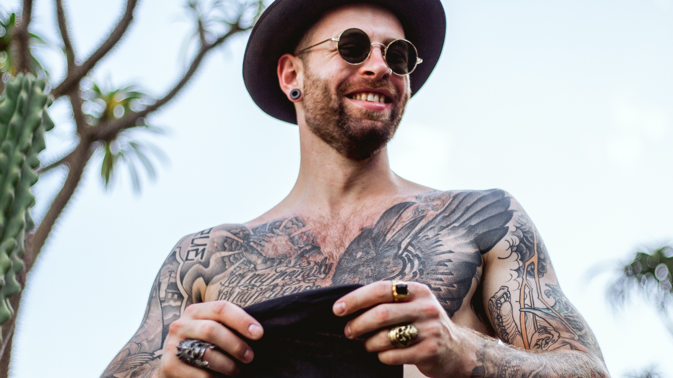 Мужчина с татуировками на теле в черной шляпе и очках