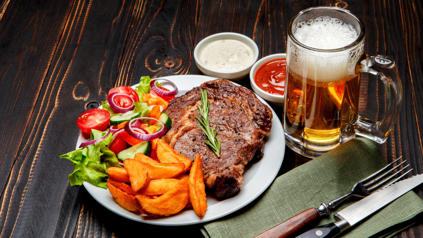 Кусок мяса на тарелке с картофелем и салатом на столе с соусом и пивом