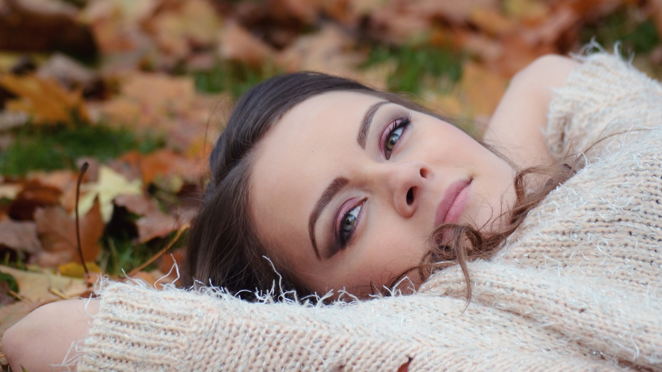 Красивая девушка в теплом свитере лежит на сухой листве 