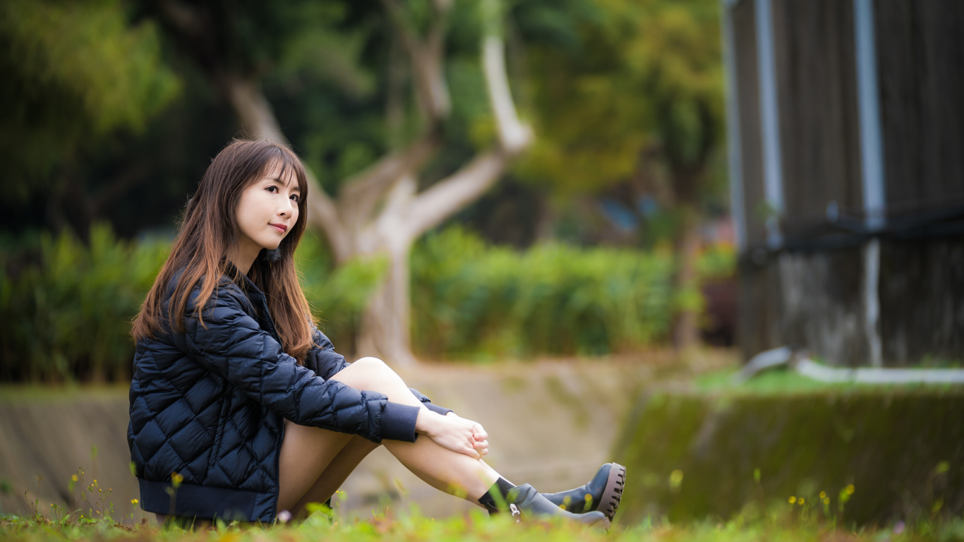 Девушка азиатка в черной куртке сидит на траве