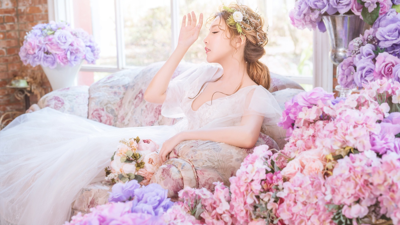 Девушка азиатка в белом платье сидит на диване в комнате с цветами