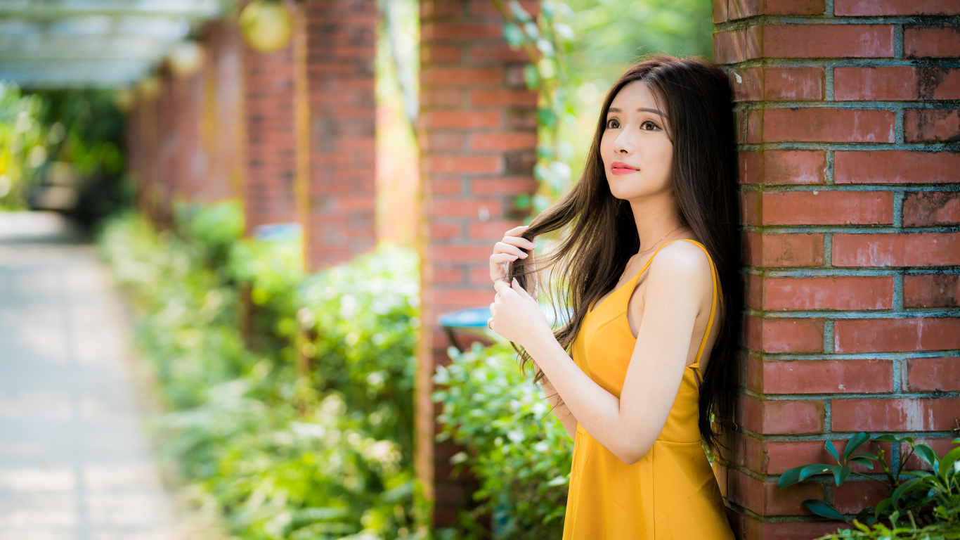 Красивая азиатка в желтом платье стоит у кирпичной колонны 