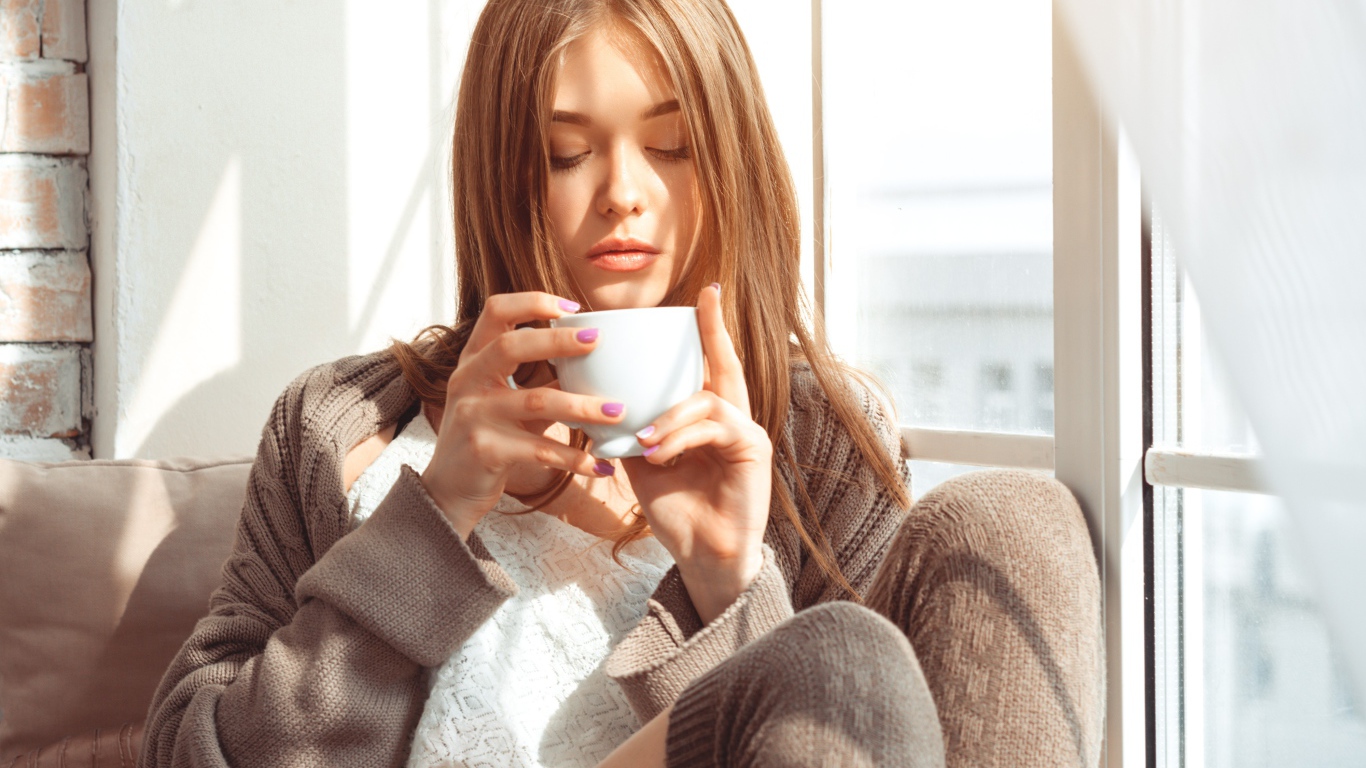 Красивая девушка в свитере пьет кофе у окна