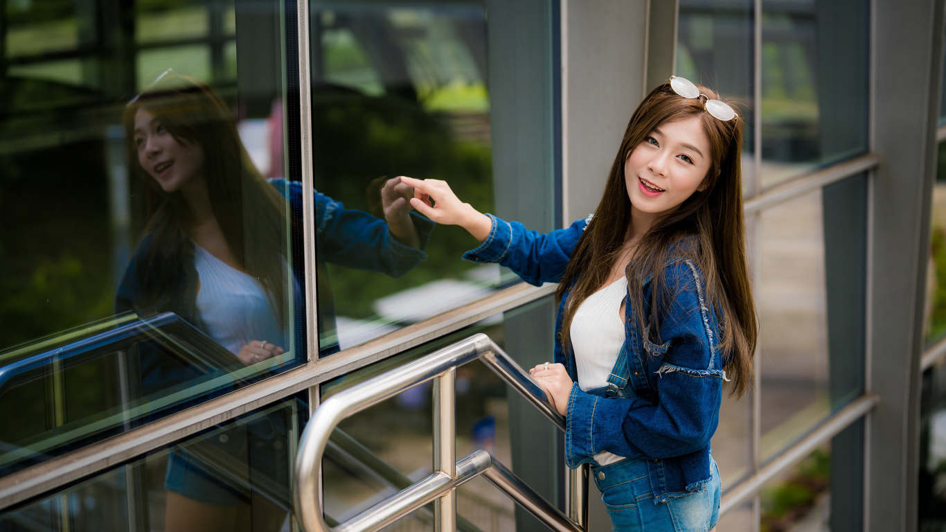 Улыбающаяся девушка в джинсовой куртке стоит у стены 