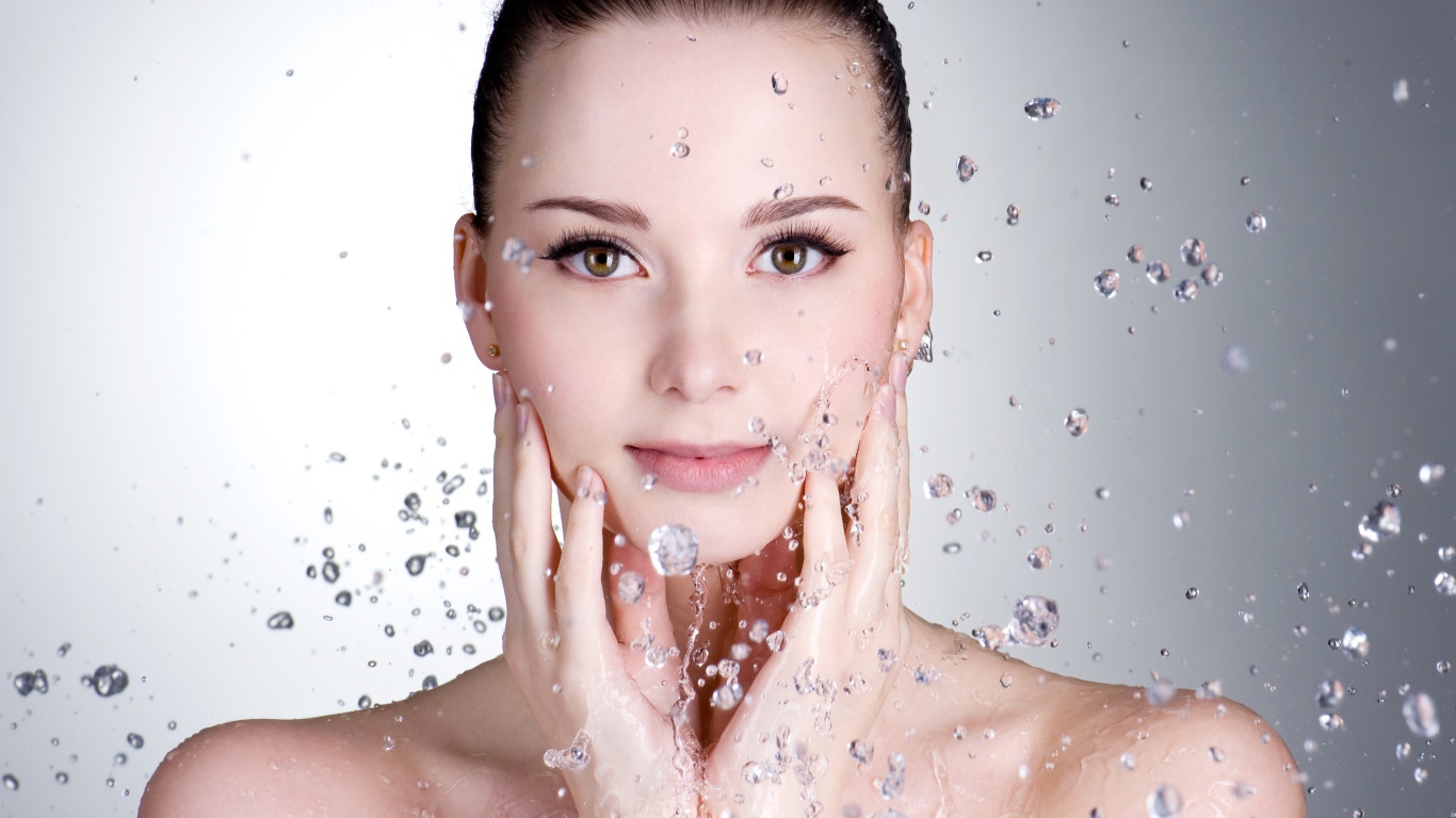 Брызги воды летят на красивое лицо молодой девушки на сером фоне