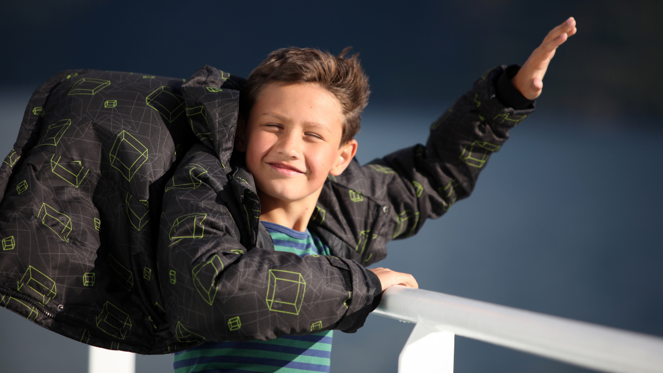 Веселый мальчик в куртке стоит на ветру в лучах солнца