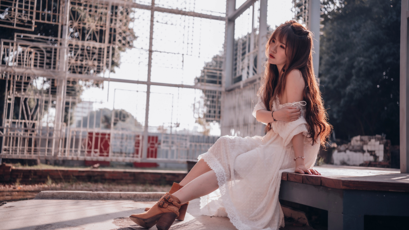 Красивая девушка азиатка в белом платье сидит на лавочке